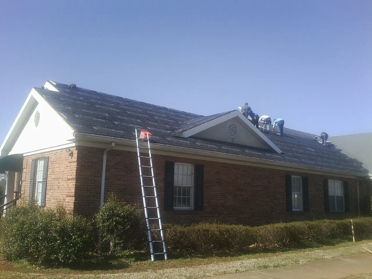 Crew tearing shingles off a brick ranch roof with a ladder set — Metal Roof Repair work by our crew in Ellenburg Center