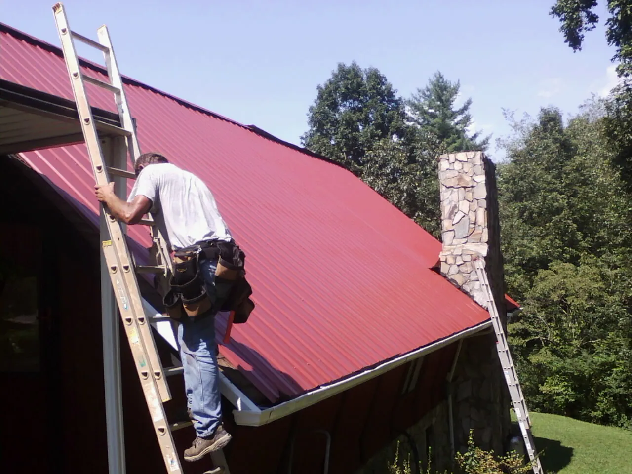 Roofer on a ladder beside a red metal roof and stone chimney — Soffit & Fascia Repair by Roofing Brothers in Ellenburg Center